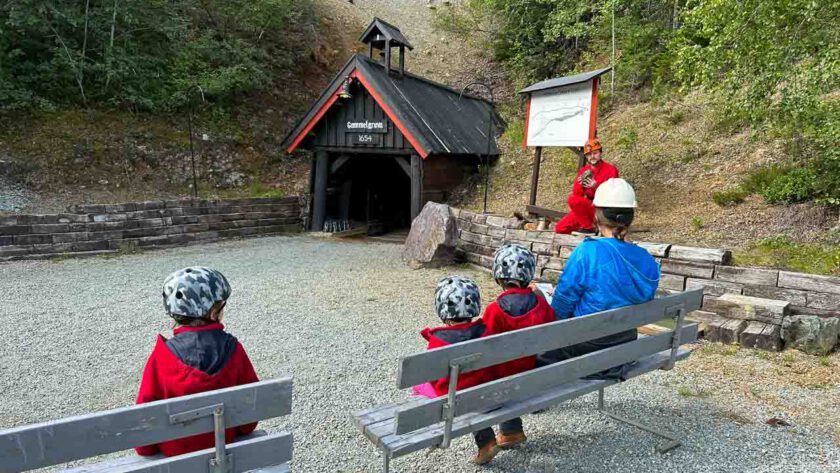 Auf Bänken vor dem Eingang des Stollens sitzen Menschen mit Helmen, die darauf warten, dass die Bergwerksführung losgeht.