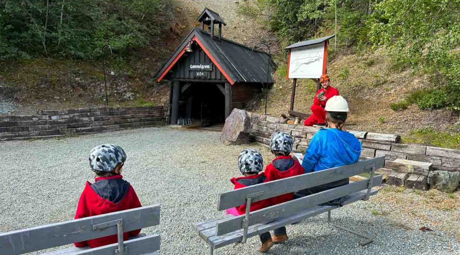 Auf Bänken vor dem Eingang des Stollens sitzen Menschen mit Helmen, die darauf warten, dass die Bergwerksführung losgeht.