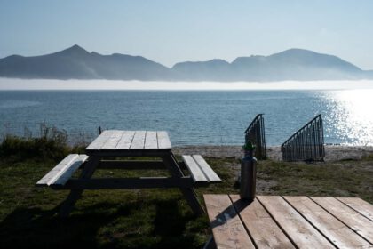 Picknicktische stehen am Meer, im Hintergrund Berge und ein leichter Nebel.