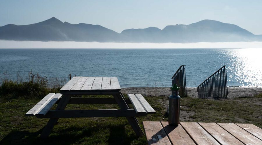 Picknicktische stehen am Meer, im Hintergrund Berge und ein leichter Nebel.