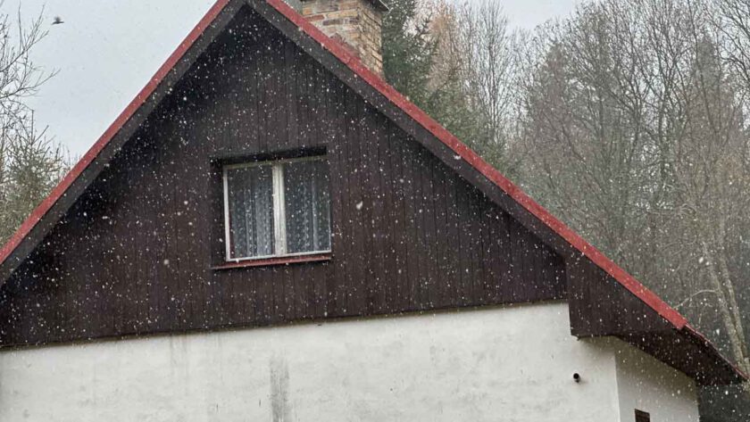 Schnee fällt vor einem Haus mit Holzfassade.